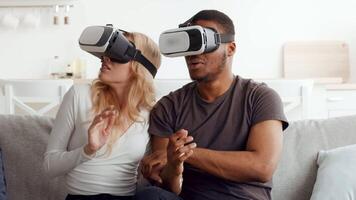 A married biracial couple experiences excitement while using virtual reality headsets in their cozy living room. They share a moment of fun and bonding in their home. video