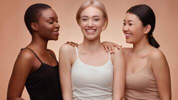 Three women of diverse backgrounds stand together, smiling and engaging in conversation. They display a sense of friendship and camaraderie against a soft, warm-colored background. video