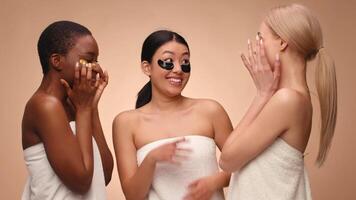 Three women wrapped in towels laugh together while applying face masks, creating a joyful and relaxing atmosphere during their spa day. They are engaged in a fun and lighthearted moment. video