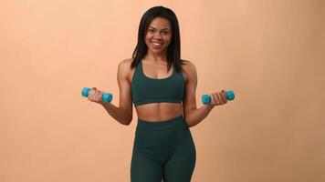 A young African American woman holds light dumbbells, smiling confidently while exercising. She embodies a healthy lifestyle in a neat studio setting designed for fitness activities. video