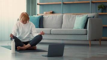 A retired Caucasian woman engages in a meditation session on a yoga mat in her cozy living room. She enjoys a moment of tranquility while following an online class. video