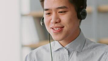 A young Asian man is focused on his work from a home office, wearing a headset. He is smiling while interacting through a telecommunication device, demonstrating remote work efficiency. video