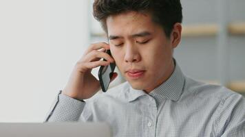 A millennial Asian man is engaged in a phone call at his home office. He appears focused and attentive, wearing a checkered shirt while seated at a desk with a laptop. video