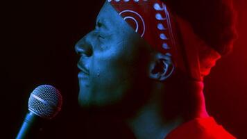 A confident millennial man stands with a microphone, passionately performing in a vibrant neon-lit setting. His cool demeanor and trendy bandana add to the captivating atmosphere. video