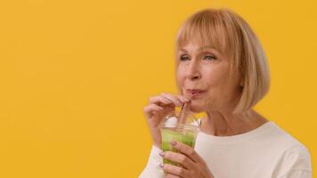 A cheerful older woman smiles while sipping a green drink with a straw, radiating joy and vitality. Vibrant yellow background enhances her lively expression and the refreshing sensation, copy space video