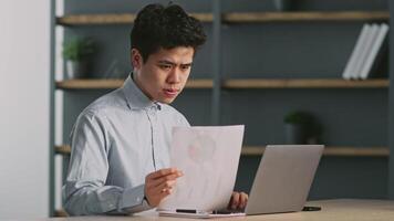 A focused Asian man is working in his home office, studying a document while sitting at a laptop. The environment is modern and organized, reflecting a productive workspace. video