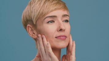 A Caucasian woman in her 50s is applying skincare products on her face. Her short hair frames her relaxed features as she focuses on pampering herself. video