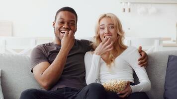 A biracial couple sits on a comfortable couch, sharing popcorn while smiling at each other. Their relaxed atmosphere radiates love and affection during their cozy evening at home. video