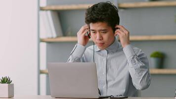 A young Asian man, dressed in a collared shirt, is seated at a desk in a contemporary home office. He is intently looking at his laptop screen while holding a headset. video