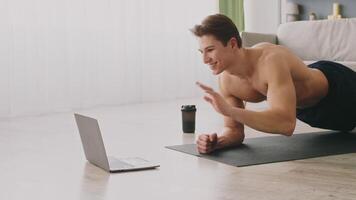 A young athletic man performs a plank exercise on a mat in his living room while focused on a laptop screen. He enjoys a health-conscious lifestyle. video