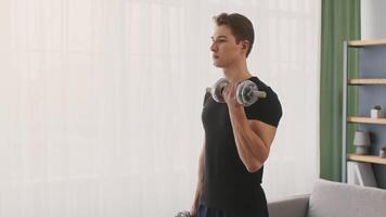 A young Caucasian man is working out at home, lifting dumbbells with focused dedication. Sunlight fills the space, highlighting his athletic build as he engages in a fitness routine. video