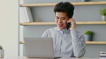 A young Asian man smiles while talking on the phone as he works at his home office. He uses a laptop on a desk in a bright, modern space with stylish decor. video