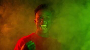 A young, handsome African American man smiles confidently against a vibrant neon background. The setting features colorful smoke effects, creating an energetic and modern atmosphere. video