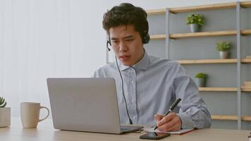 A young Asian man is engaged in remote work at his home office. He wears a headset while typing on his laptop and notes on a notepad, focused on his tasks. video
