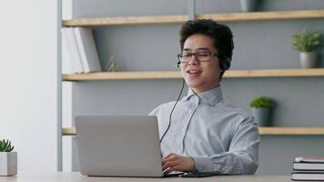 A young Asian man engages in a virtual meeting from his home office. He is using a laptop and headset, appearing focused and professional surrounded by minimal decor. video