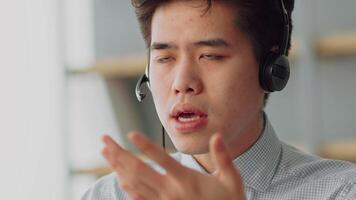 A young Asian man engages in an online work call from his stylish home office. He wears a headset and focuses on a computer screen, demonstrating a professional remote working environment. video