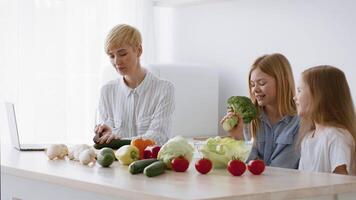A mother and her two daughters enjoy a cooking session at home, reviewing recipes on a laptop. Fresh vegetables are spread across the table as they prepare healthy meals together. video
