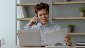 A young Asian man sits at his home office desk while talking on the phone and reviewing documents. The casual yet focused atmosphere highlights remote work dynamics in a modern setting. video
