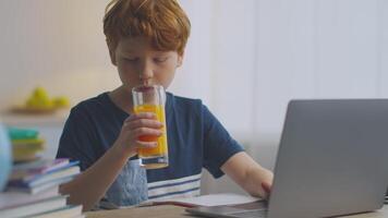 A preteen boy with red hair focuses intently on his laptop while studying at home. He enjoys a glass of orange juice, surrounded by books and learning materials. video