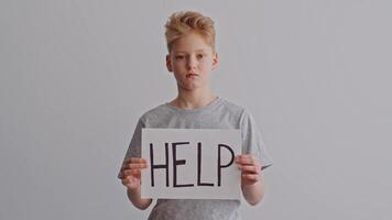 A young boy stands against a plain backdrop with a serious expression, holding a large sign that reads HELP. His demeanor suggests distress and a pressing need for support. video