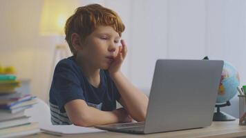 A young boy with ginger hair sits at a desk, studying remotely. He appears frustrated while covering his face with one hand, surrounded by books and a globe in a well-lit space. video