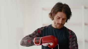 A young Caucasian man is getting ready for a boxing workout at home, adjusting his red gloves and focused on his exercise routine. The environment is bright and motivating, copy space video