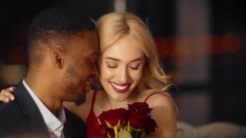 A young, diverse couple shares an affectionate moment during a dinner date at an upscale restaurant. The woman holds a bouquet of roses while smiling at her partner, who wears a suit. video