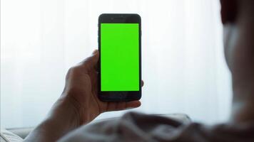 A young African American man sits comfortably at home, deeply focused on his smartphone. The device displays a bright green screen, indicating possible content creation or app usage. video