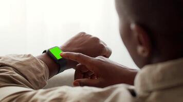 A young African American man sits comfortably at home, engaging with his smartwatch. He is focused on the device's screen, indicating a moment of curiosity and interaction with technology. video
