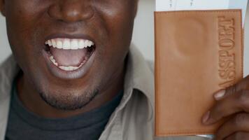 A young African American man smiles widely while holding a passport and travel documents. He appears excited about future travel opportunities in a comfortable home setting. video