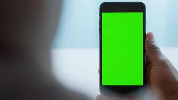 A young African American man sits comfortably in his home, focused on his smartphone, displaying a vibrant green screen. He explores technology and reflects on digital life. video
