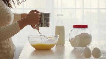 A woman is engaged in cooking and baking at home, sifting flour into a bowl of eggs while surrounded by ingredients like milk and sugar. The kitchen is bright and inviting. video