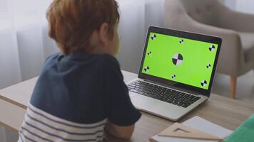 A boy with ginger hair focuses intently on a laptop screen while studying at home. The bright, inviting study area reflects a positive remote learning environment during the day. video