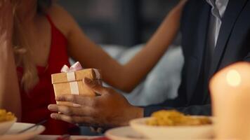 A diverse couple sits closely together at a table in a cozy restaurant, sharing a romantic dinner. The woman hands a beautifully wrapped gift to her partner, filled with affection. video