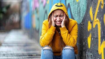 A young person wearing a yellow jacket sits on the ground against a colorful wall, expressing feelings of sadness and frustration. The setting reflects an urban environment, emphasizing emotions. video
