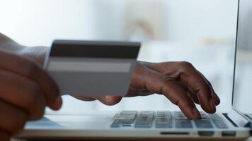 A young African American man sits at home, focused on his laptop. He is holding a credit card in one hand while typing with the other, engaged in an online transaction or shopping. video