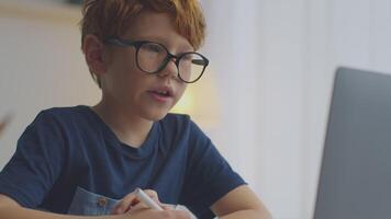 A preteen boy with ginger hair is focused on his online lesson while studying at home. He is wearing large glasses, taking notes, and interacting with a laptop in a bright setting. video