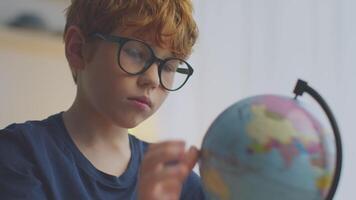 A boy with ginger hair and glasses is focused on a globe while engaging in remote learning. He is exploring different countries and honing his geography skills from his study area. video