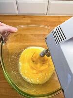 hand holds a mixing bowl while an electric mixer whips egg yolks in kitchen . High quality photo
