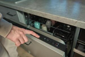 Hand pressing start button on dishwasher control panel with open door. Woman using modern home appliance, choosing automatic program on built in dishwasher for cleaning dirty dishes photo