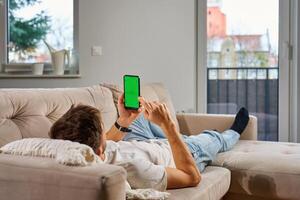 Man in casual clothes lies on beige couch, holding smartphone with green screen, relaxing in modern living room. Male resting on sofa and watching online on mobile phone with chroma key display photo