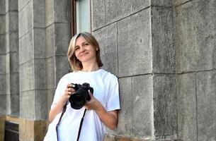 This is a portrait of a woman holding a DSLR camera with a stone wall of a building as a backdrop. She is looking to her left and has a soft smile. photo