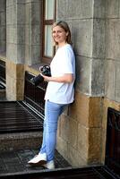 The photograph shows a woman in a simple outfit of a white shirt and light blue jeans, standing against a backdrop of an ornate building with grand columns. photo