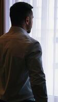 Man in hotel near the window. Young businessman in white shirt standing near the window and viewing through it in the hotel room. Back view. Vertical video