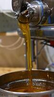 Fresh amber honey quickly flowing from metal barrel faucet. Organic product being poured onto sieve for filtration. Blurred backdrop. Vertical video