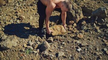 Athletic man exercises with stones. Strong sportsman taking heavy stone and throwing it over the head behind the back on rocky background. Slow motion. video