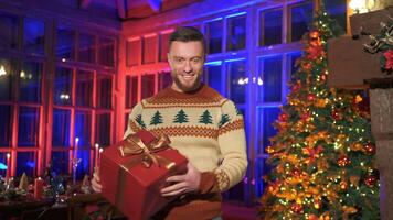 Smiling man offering new year present. Portrait of a handsome man in knitted Christmas sweater holding a box with gift on Christmas decorated room background. video
