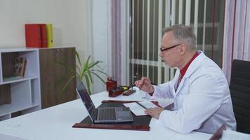 Doctor listens to a patient through the remote webcam on laptop screen. Professional male doctor in hospital office talking to a client online by call on computer. Profile view. video