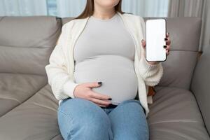 Close-up photo of pregnant woman holding and showing mobile phone with empty display at home. Mockup image with copy space for app or interface customization