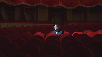 Rich man among rows of empty red chairs. Businessman in medical mask sitting in comfortable chairs alone on dark theater background. Pandemic in the world. video